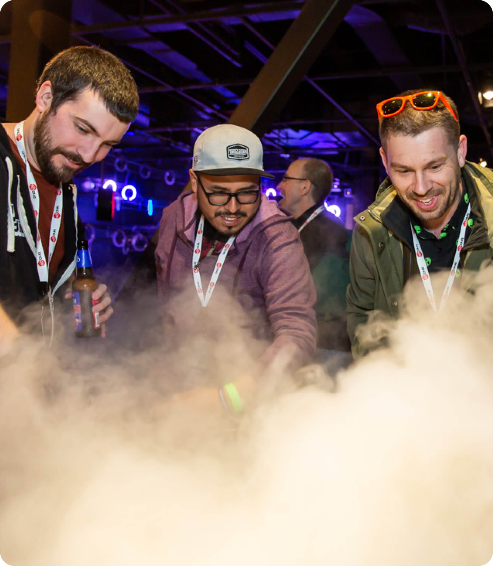 Three men wearing event lanyards stand around a table with fog or smoke, with one holding a bottle; an indoor event space is in the background.