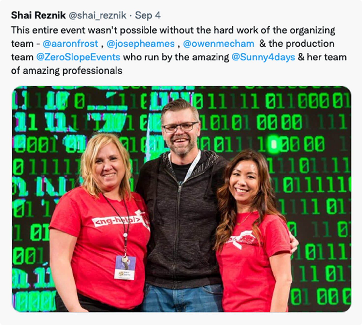 Three people stand smiling in front of a digital green binary code background; two women in red shirts flank a man in a gray shirt.