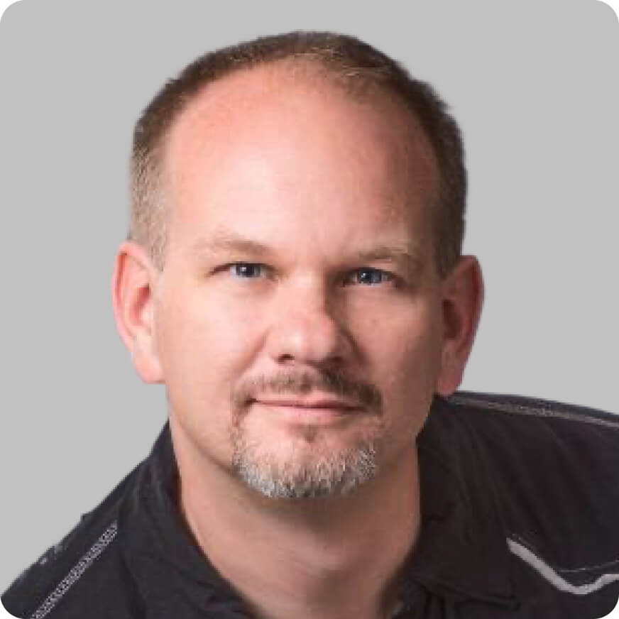 A middle-aged man with short light hair and a trimmed beard, wearing a dark shirt, poses against a plain gray background—capturing the confident demeanor of a Vibecoding con AI expert.
