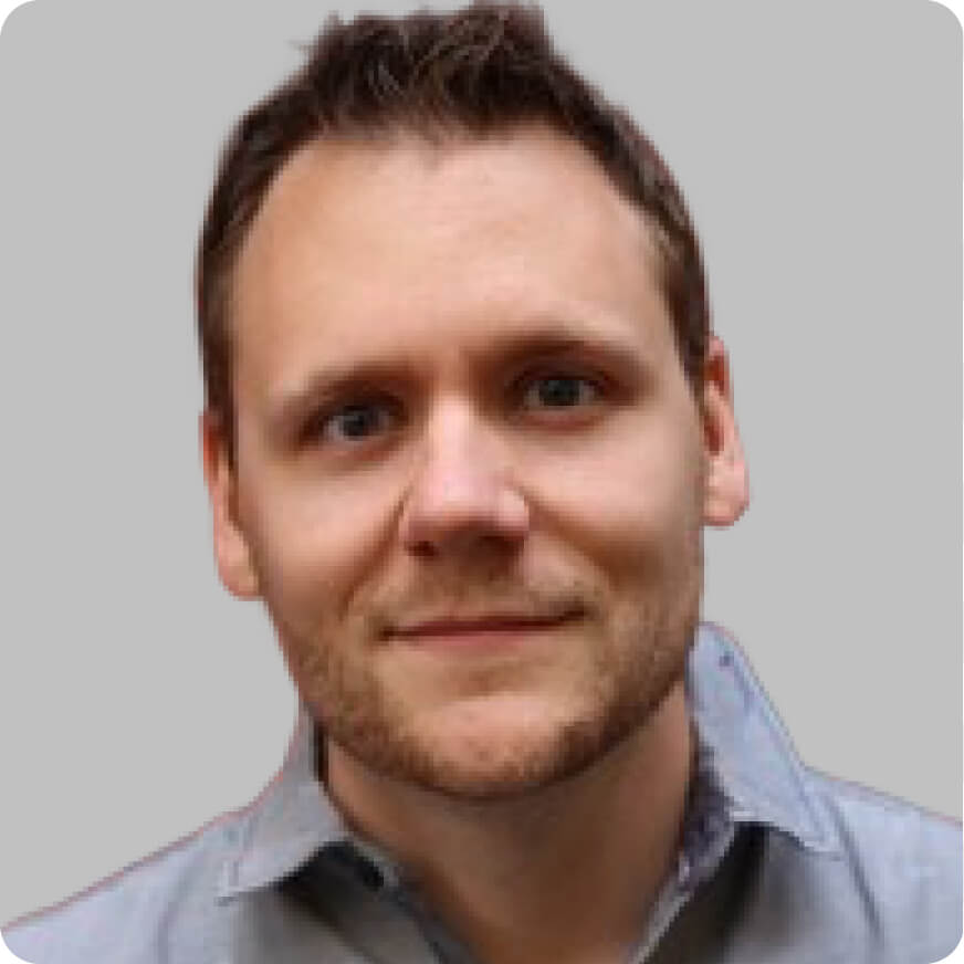 A man with short brown hair, light skin, and facial hair, wearing a light grey collared shirt, faces the camera against a plain grey background—portraying the look of a Vibecoding con AI expert.