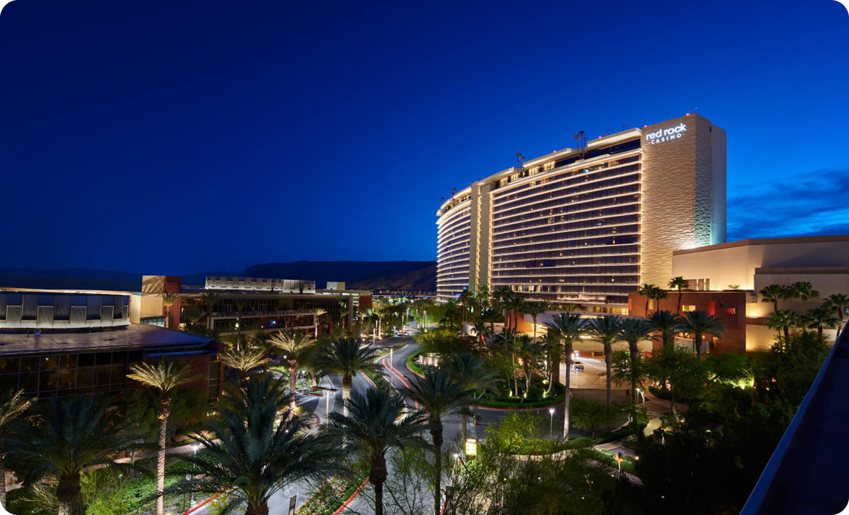A large, well-lit hotel with the sign "Red Rock Resort" is surrounded by palm trees and a driveway at dusk under a dark blue sky.