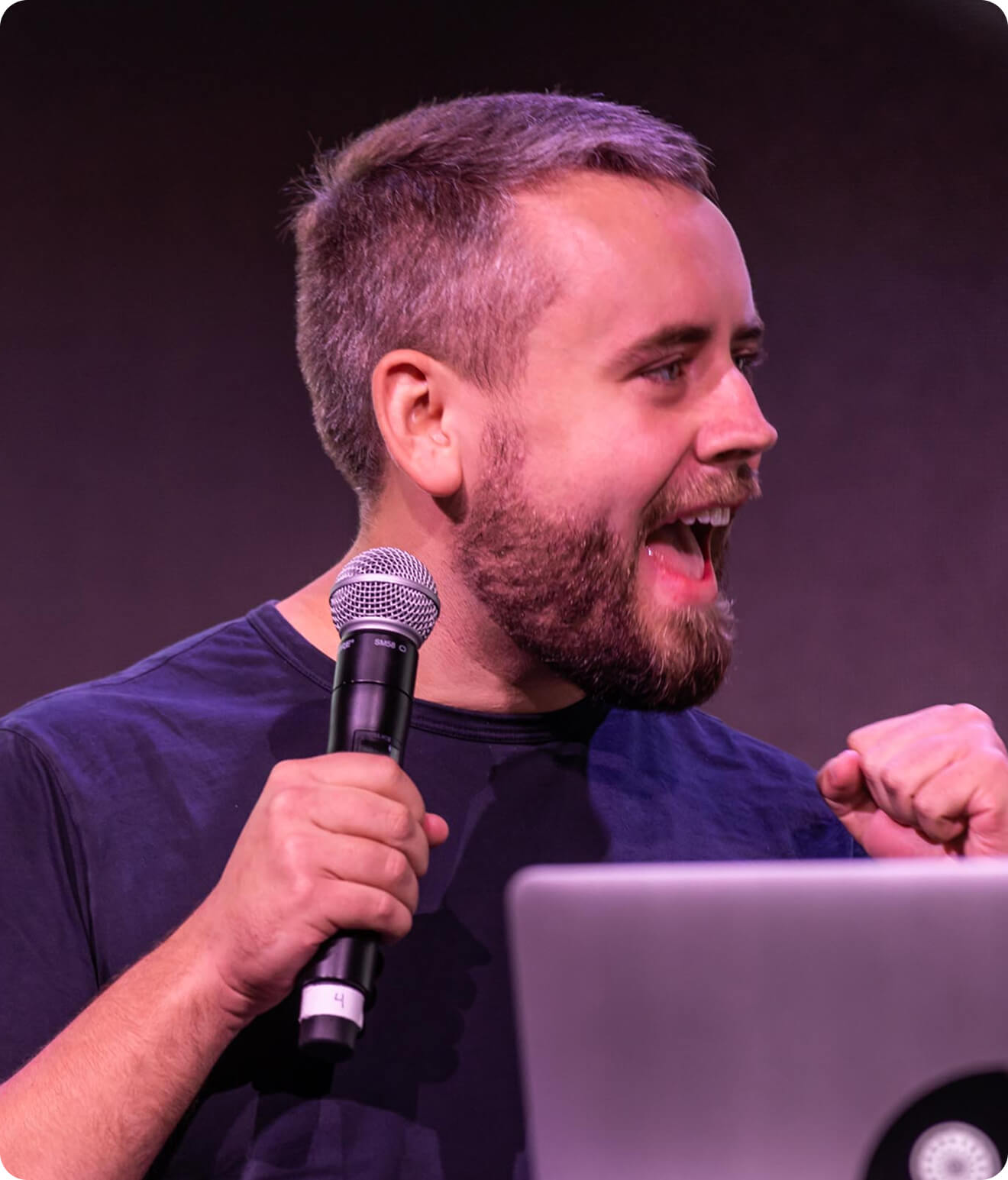 A man with a beard holds a microphone and gestures with his fist, appearing to speak enthusiastically on stage next to a laptop.