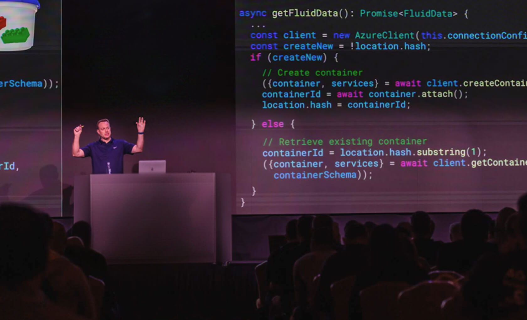 A speaker stands at a podium presenting to an audience, with a large screen behind him displaying programming code.