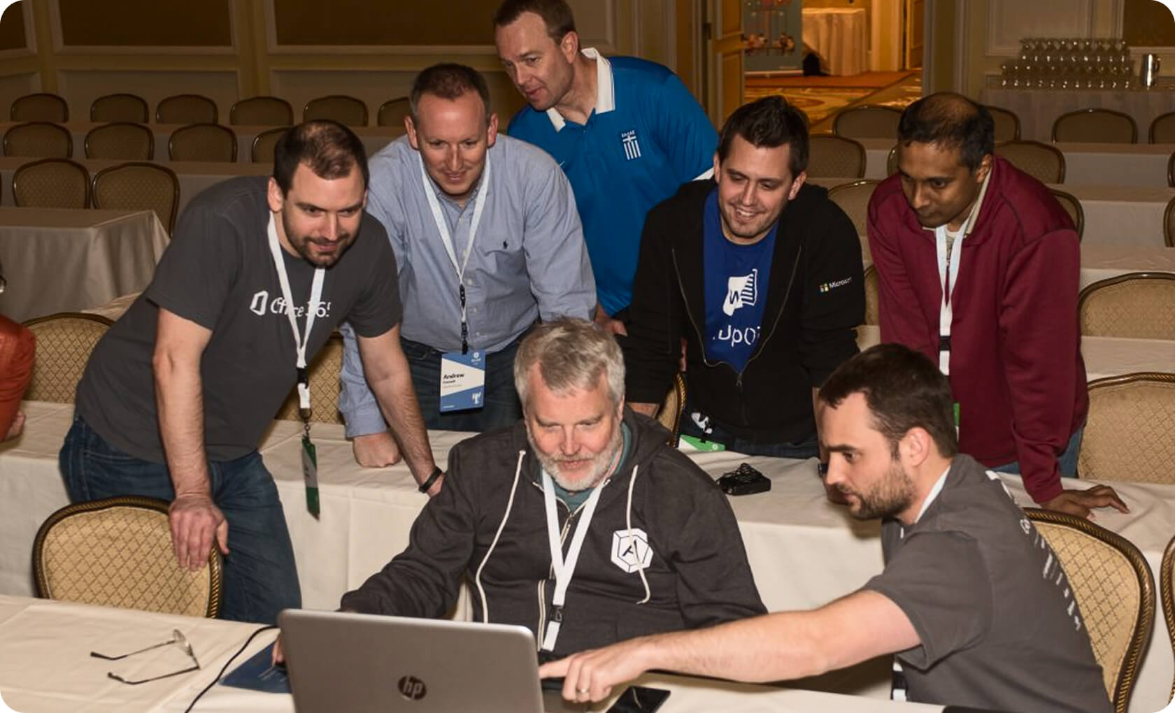 Six men are gathered around a laptop in a conference room, with one man seated typing while the others stand or crouch closely watching the screen.