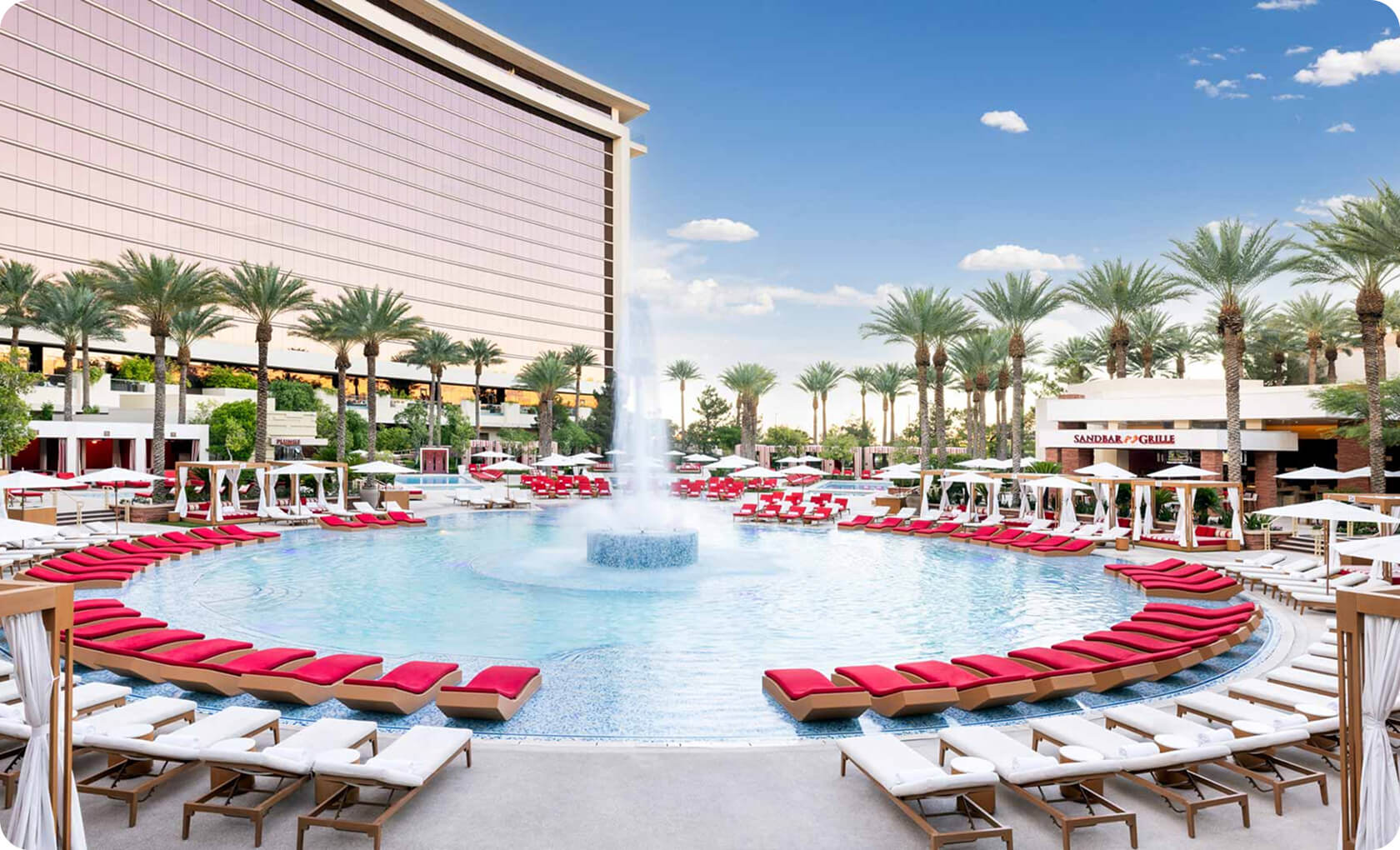 Circular outdoor pool with a central fountain, surrounded by red and white lounge chairs, palm trees, and resort buildings under a blue sky.