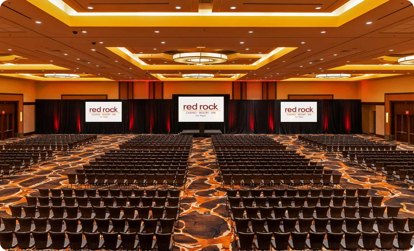 Large conference room with rows of empty chairs facing two projection screens displaying "Red Rock Casino Resort Spa" logos under warm ceiling lights.