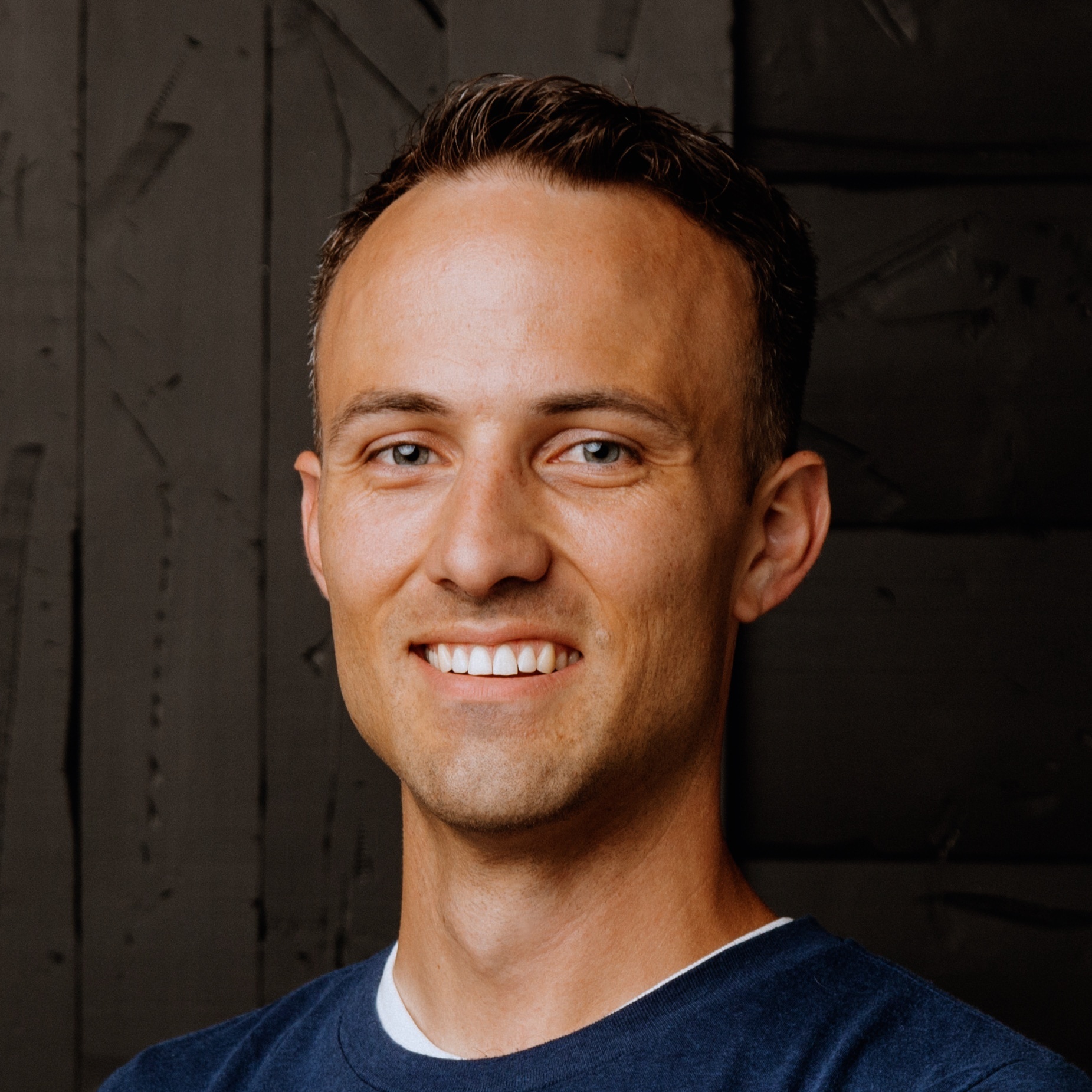 Smiling man with short brown hair wearing a navy blue shirt stands in front of a dark textured wall.