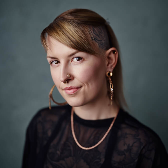 Person with nose and ear piercings, light blonde hair partially shaved, and visible tattoos, wearing a sheer black top and gold jewelry, poses against a plain green background—bringing creative energy reminiscent of dev AI tools to their effortlessly bold look.