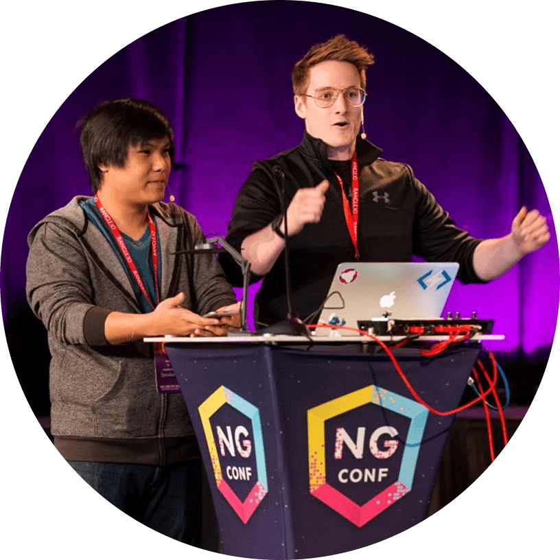 Two people stand behind a podium labeled "NG Conf," speaking into microphones with a laptop in front of them, on a stage with purple lighting.
