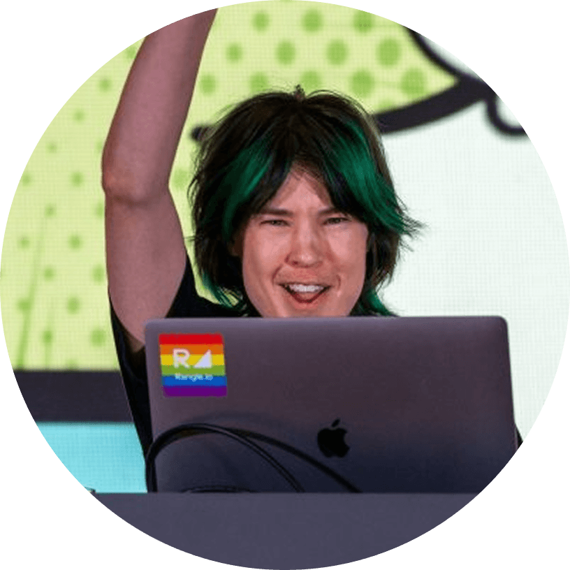 Person with green and black hair raises one arm while smiling, sitting behind a laptop with a rainbow pride sticker, in front of a brightly colored background.