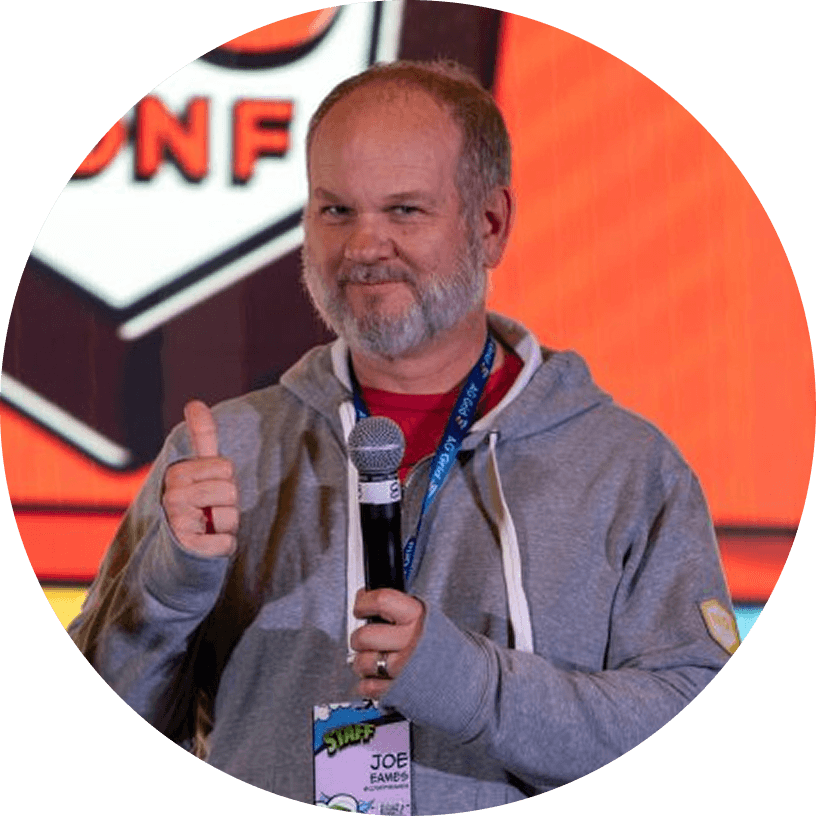 A man in a gray hoodie holds a microphone and gives a thumbs up on stage. A conference badge hangs around his neck. An orange and white event sign is in the background.