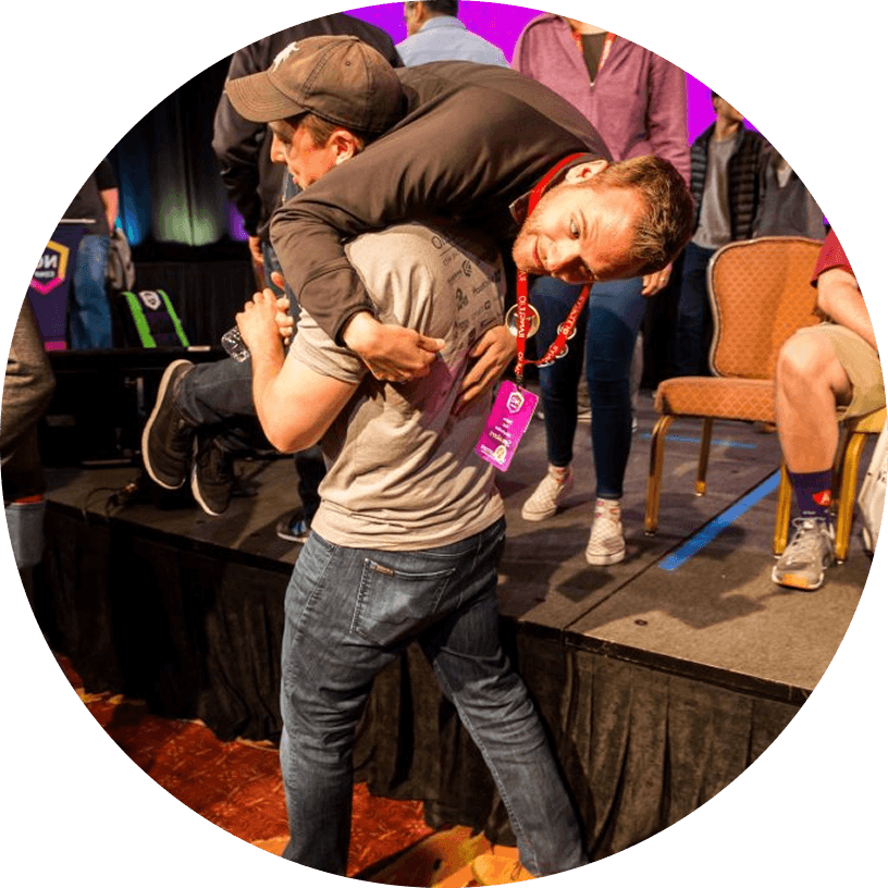 A man carries another man over his shoulders at an indoor event. People sit and stand in the background, and event badges are visible.