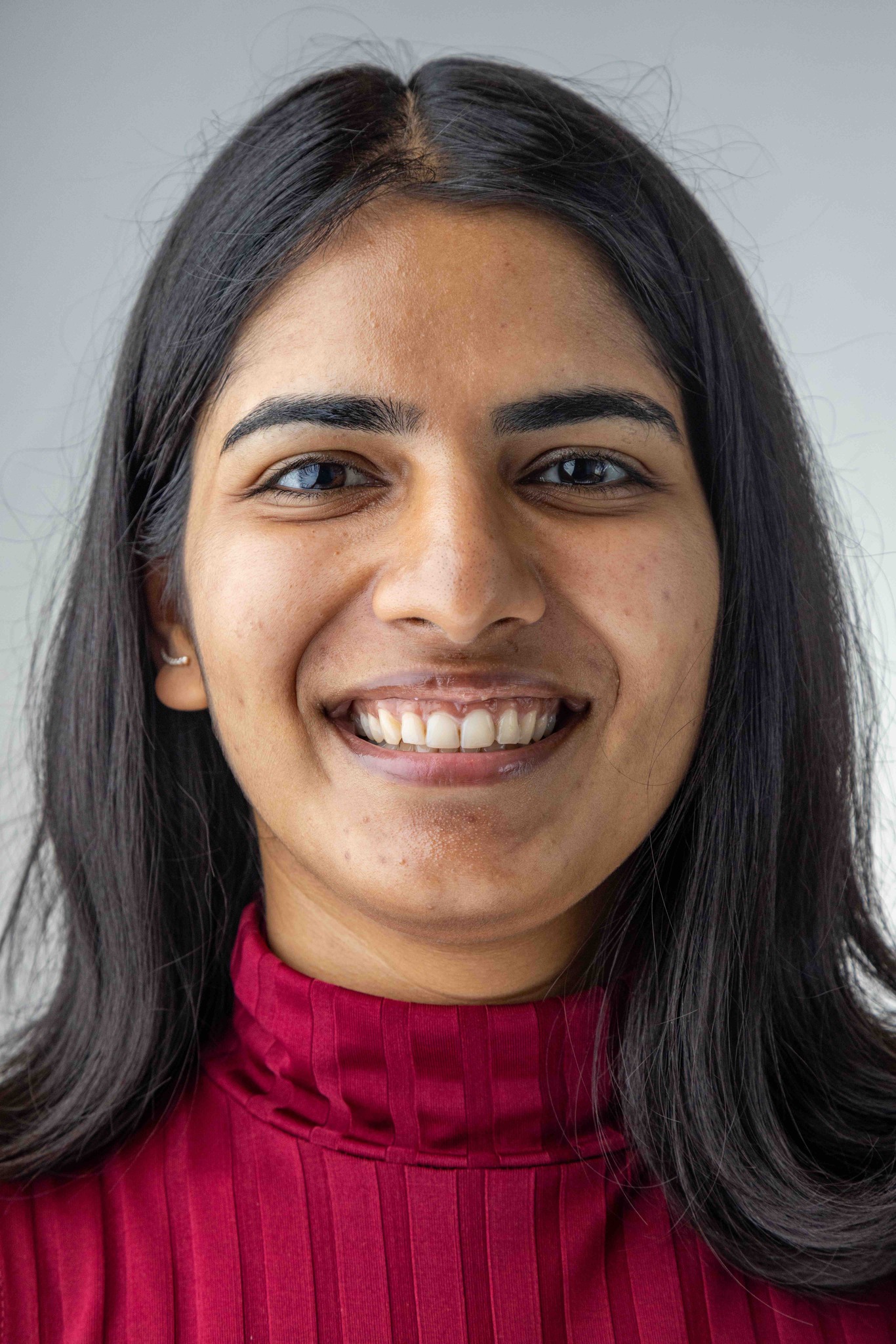 A woman with long dark hair, wearing a red high-neck top, smiles at the camera against a plain light background.