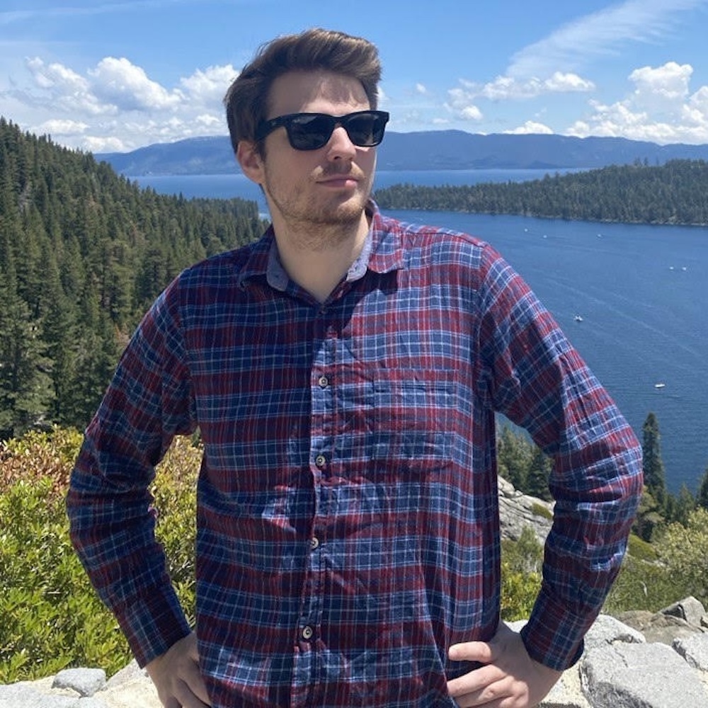 A person wearing sunglasses and a plaid shirt stands outdoors with a lake, trees, and mountains in the background under a partly cloudy sky.