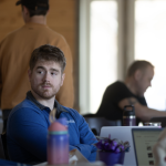 A man in a blue shirt sits at a table indoors, looking to the side. Other people are nearby, some using laptops. The background is softly lit with natural light.