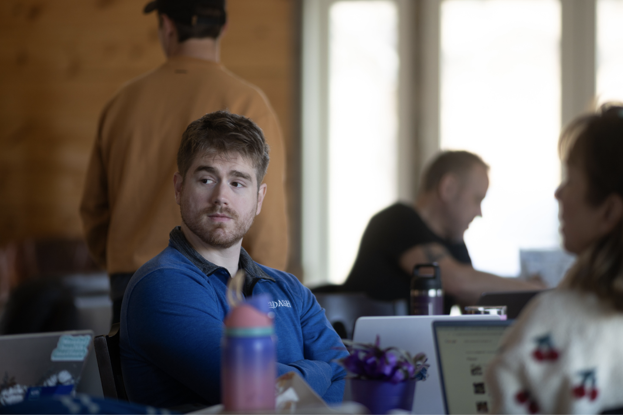 A man in a blue shirt sits at a table indoors, looking to the side. Other people are nearby, some using laptops. The background is softly lit with natural light.