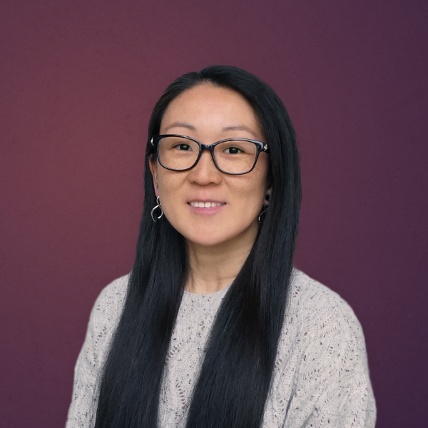 A woman with long black hair and glasses wears a light knit sweater and hoop earrings, posing against a plain purple background.