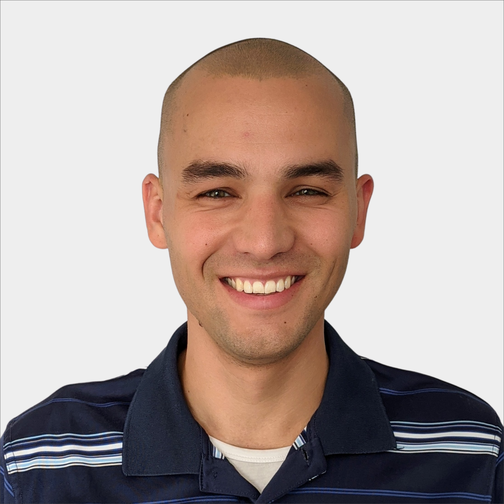 A person with a shaved head and dark eyebrows smiles while wearing a navy blue collared shirt with white stripes, in front of a plain light background.