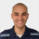 A person with a shaved head and dark eyebrows smiles while wearing a navy blue collared shirt with white stripes, in front of a plain light background.