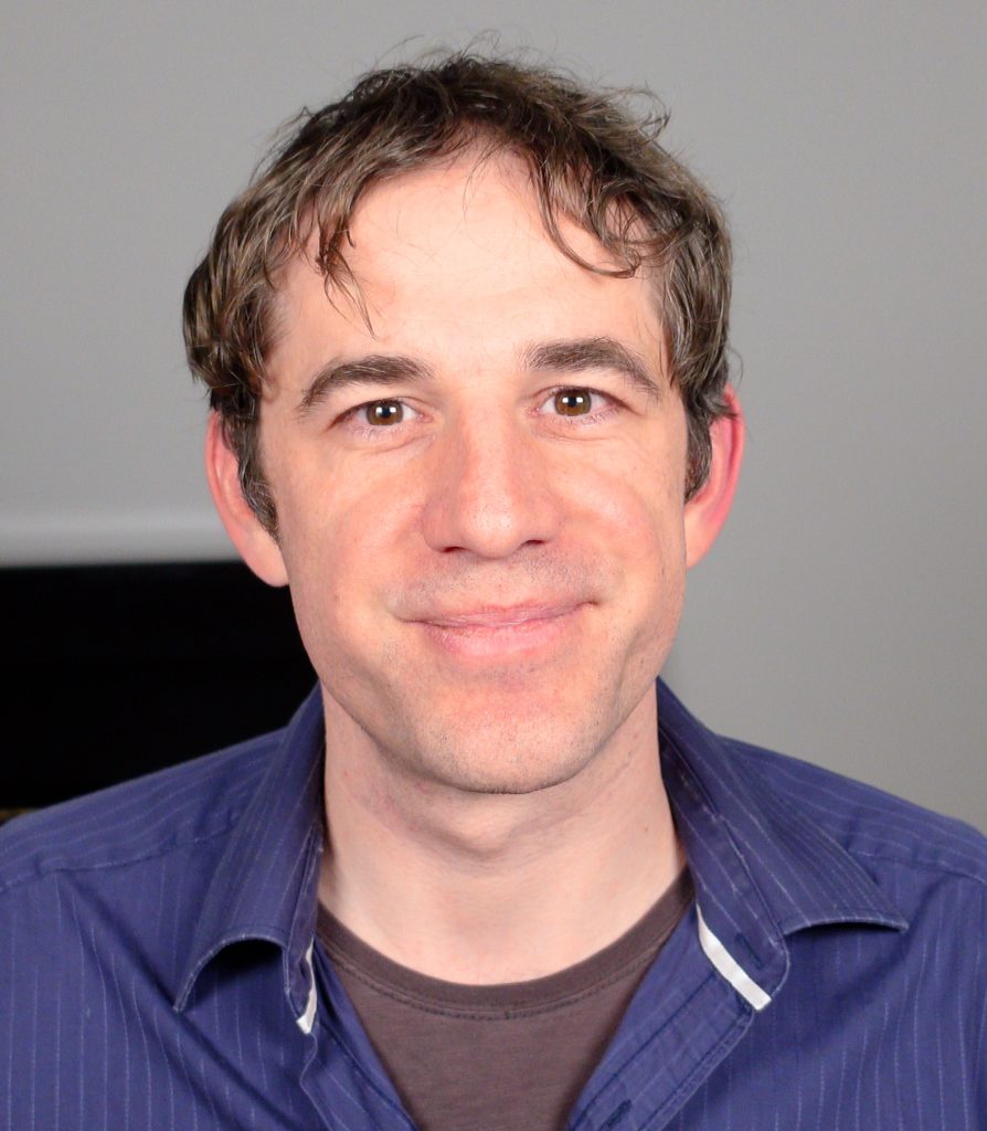 A man with short brown hair and a blue collared shirt smiles at the camera against a plain background.