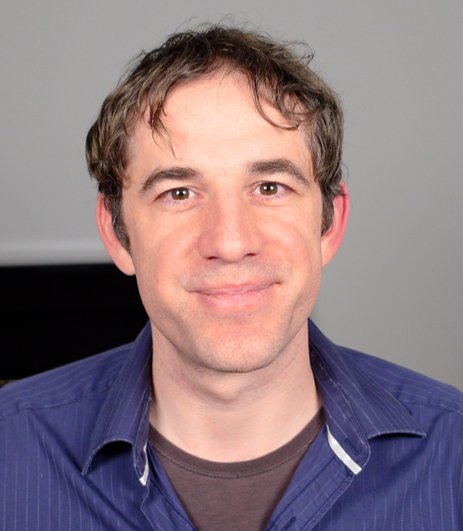 A man with short brown hair and a blue collared shirt smiles at the camera against a plain background.