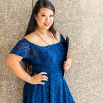 Woman with long dark hair in a blue lace dress, wearing a pearl necklace, stands against a stone wall holding a closed laptop.