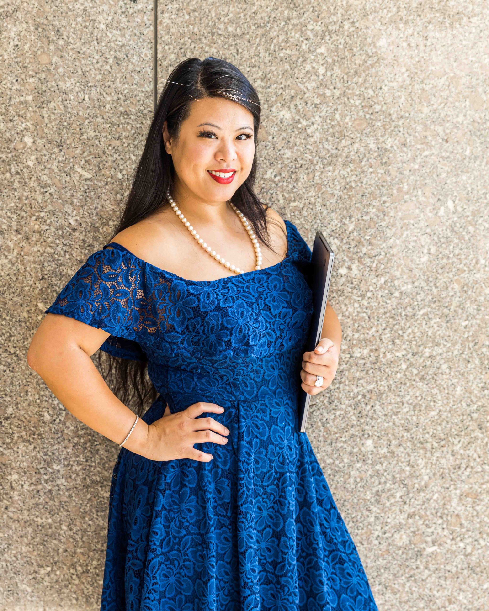 Woman with long dark hair in a blue lace dress, wearing a pearl necklace, stands against a stone wall holding a closed laptop.