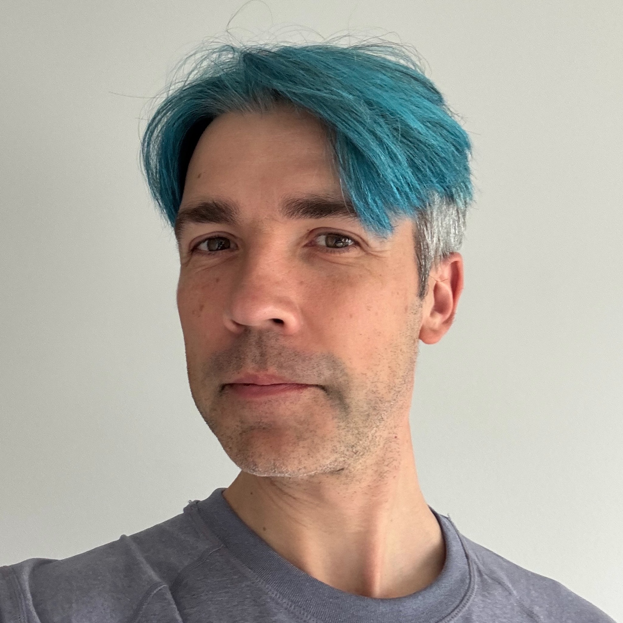 A person with short blue and gray hair, wearing a gray shirt, stands in front of a plain light-colored wall, looking slightly to the side.
