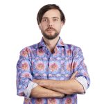 Man with a beard and mustache stands with arms crossed, wearing a colorful, patterned shirt against a plain white background.