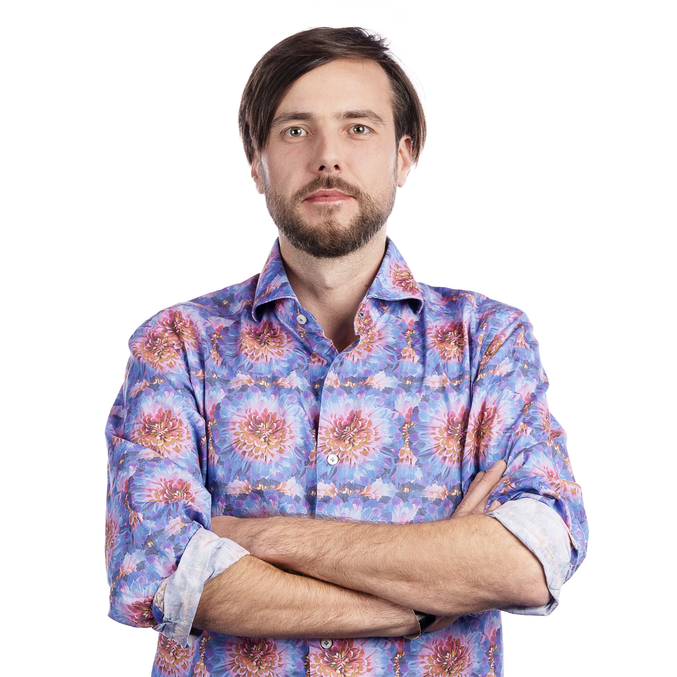 Man with a beard and mustache stands with arms crossed, wearing a colorful, patterned shirt against a plain white background.