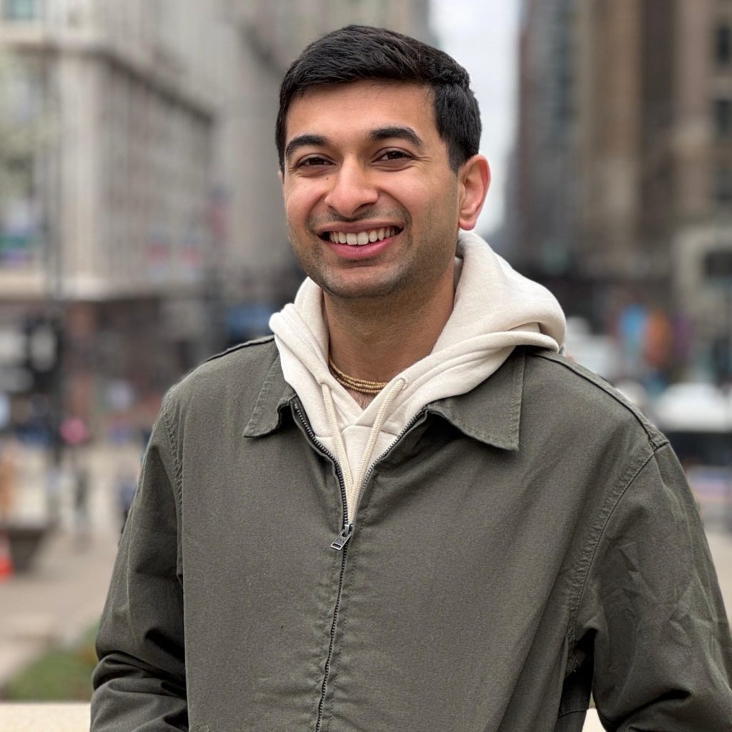 A smiling man wearing a beige hoodie and olive green jacket stands on a city street.